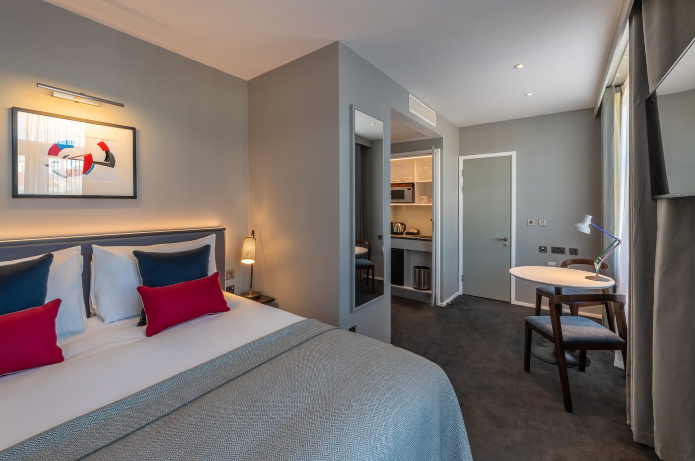 A modern hotel room with a neatly made double bed, colorful pillows, gray bedding, a small dining area with chairs, kitchenette, wall art, large window with curtains, and neutral-colored walls and carpet.