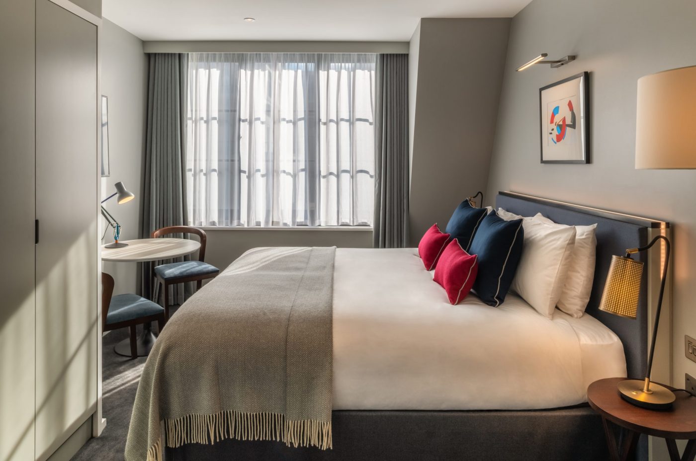 Modern hotel room with a large bed, white linens, and colorful pillows. A small round table with two chairs sits by a window with sheer curtains. Minimalist decor includes a framed abstract artwork above the bed.