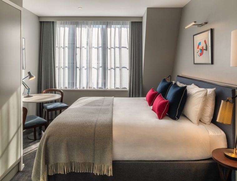 Modern hotel room with a large bed, white linens, and colorful pillows. A small round table with two chairs sits by a window with sheer curtains. Minimalist decor includes a framed abstract artwork above the bed.