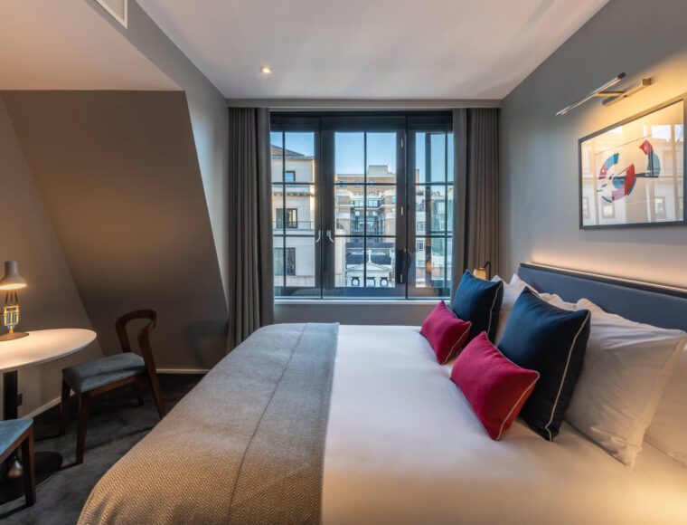 A modern hotel room with a large bed featuring white linens and colorful pillows, a small table with two chairs, a window with city views, and a framed abstract artwork on the gray wall.