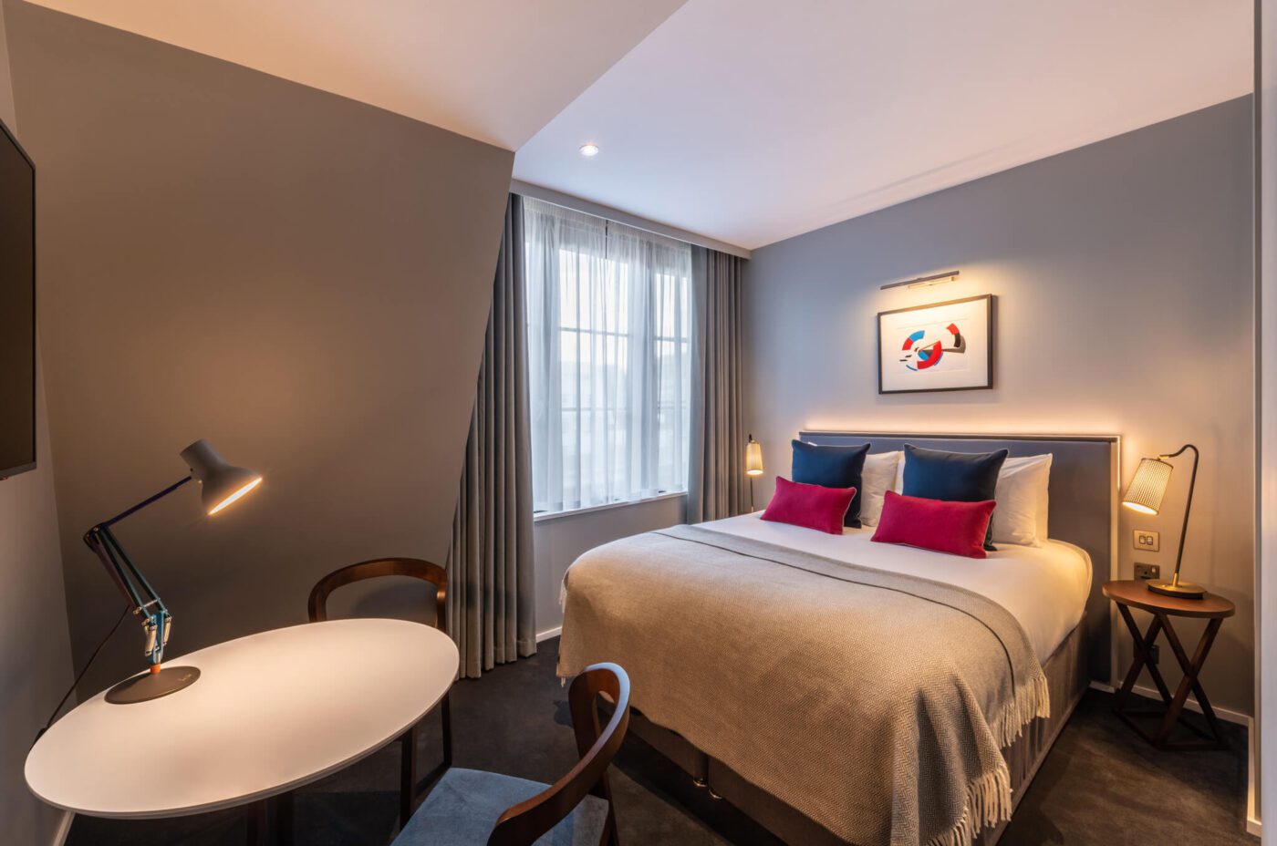 A modern hotel room with a large bed, gray walls, colorful pillows, a round table with two chairs, a desk lamp, a window with curtains, and framed artwork above the bed.