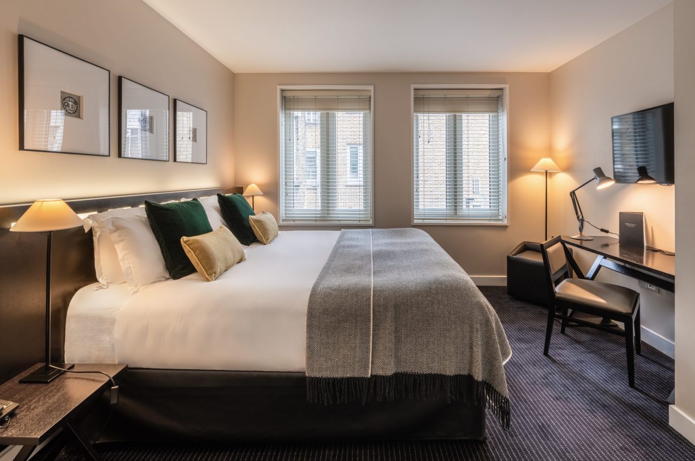A modern hotel room with a large double bed, neatly made with white linens and decorative pillows. The room features two windows, wall art, a flat-screen TV, a desk with a lamp, and warm ambient lighting from floor lamps.