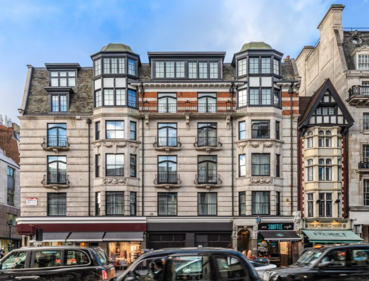 A street view of a London building with a classic architectural design featuring multiple bays and large windows. The ground floor houses several shops, including a restaurant and a barber shop. Black taxis and cars are visible on the busy street in front, reminiscent of areas near The Resident Covent Garden.