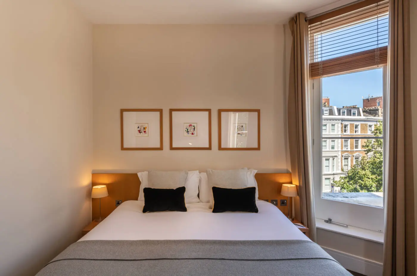 A cozy bedroom at The Resident Kensington features a neatly made bed with white and grey linens, accompanied by black and white pillows. Three framed artworks hang above the bed. The room is well-lit by natural light from a large window with beige curtains, offering a view of buildings outside.