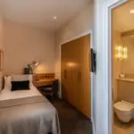 A cozy hotel room at The Resident Kensington featuring a single bed with a beige blanket and pillows, a wooden desk with a lamp and chair, large wooden wardrobe, and an en-suite bathroom with a sink, toilet, and towel rack. The space is warmly lit and simply decorated.
