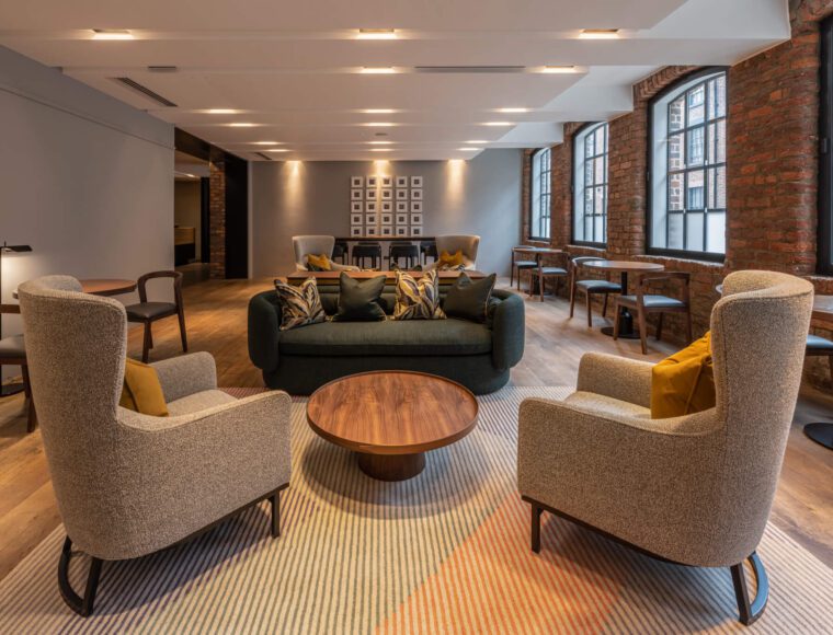 A modern lounge area at The Resident Liverpool features two cushioned armchairs and a dark green sofa arranged around a circular wooden coffee table on a striped rug. The space boasts exposed brick walls, large windows, and wooden floors, with several small tables and chairs in the background.