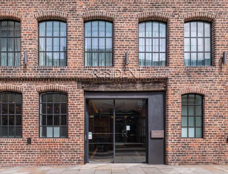 A brick building facade with large windows, featuring an arched main entrance under a wooden beam. Above the entrance, the words 