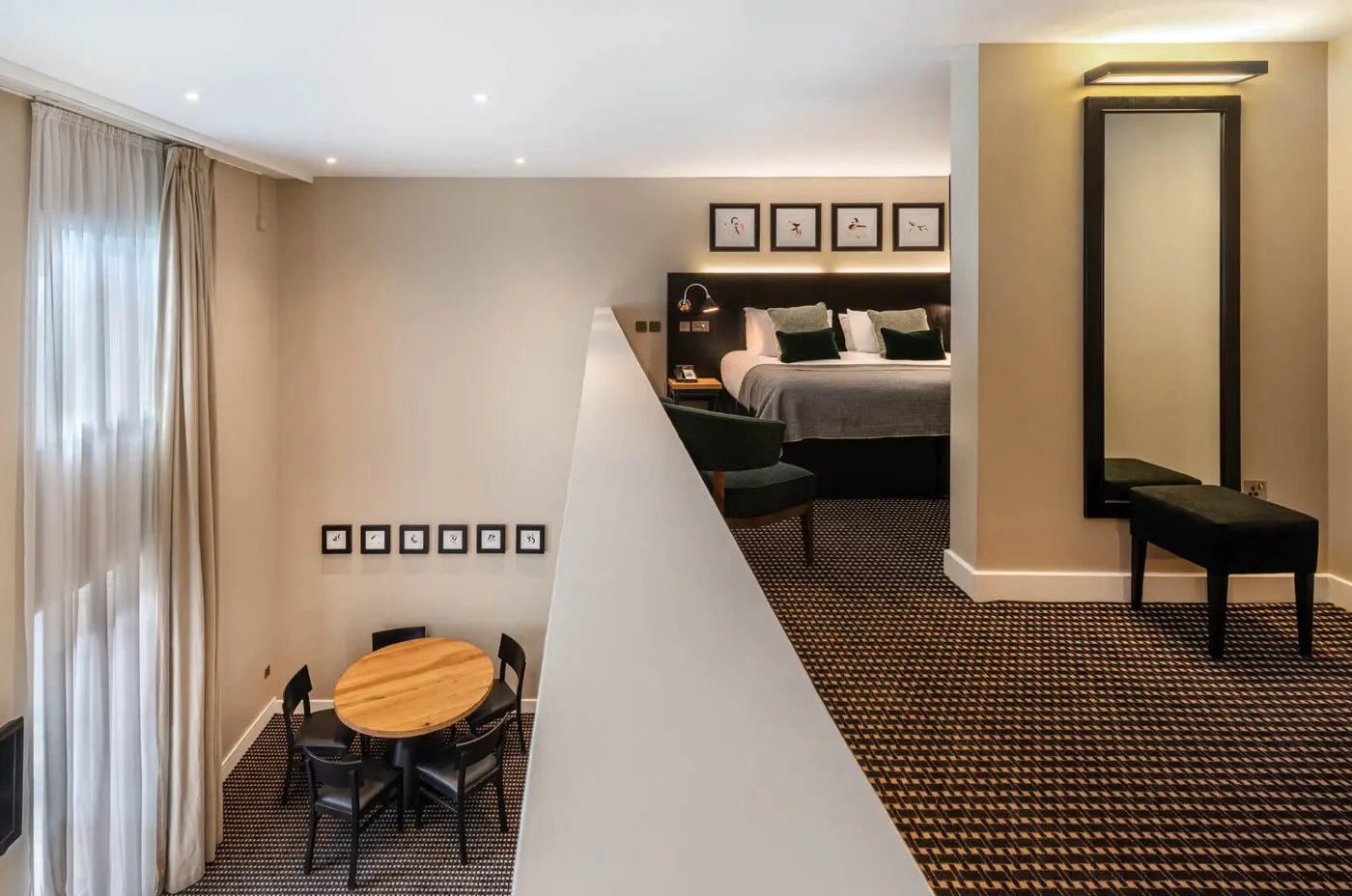 A split-level room at The Resident's Liverpool Hotel features a lower level with a round dining table and four black chairs. The upper level has a bed, framed art above it, a large mirror, and cozy seating. Both levels have beige walls and patterned flooring, with ample natural light from tall windows.