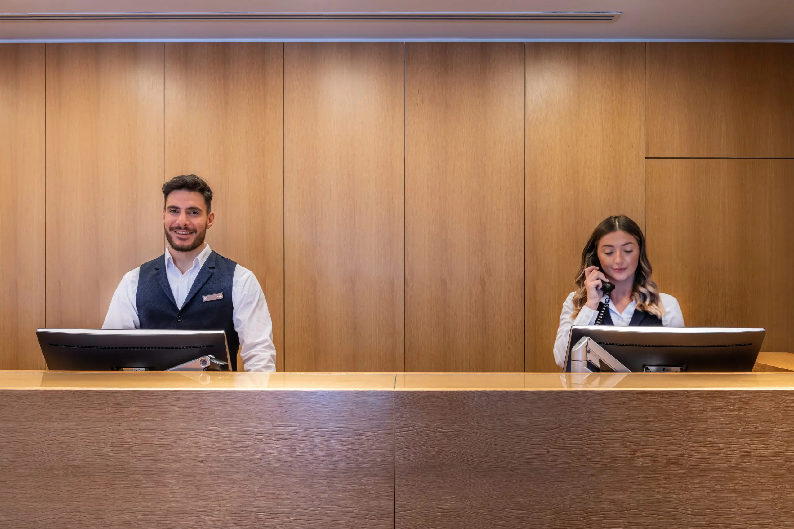 Two hotel receptionists stand behind a wooden counter at The Resident Liverpool. The male receptionist on the left smiles at the camera, while the female receptionist on the right speaks on the phone. Both have computers in front of them and are dressed in professional attire.