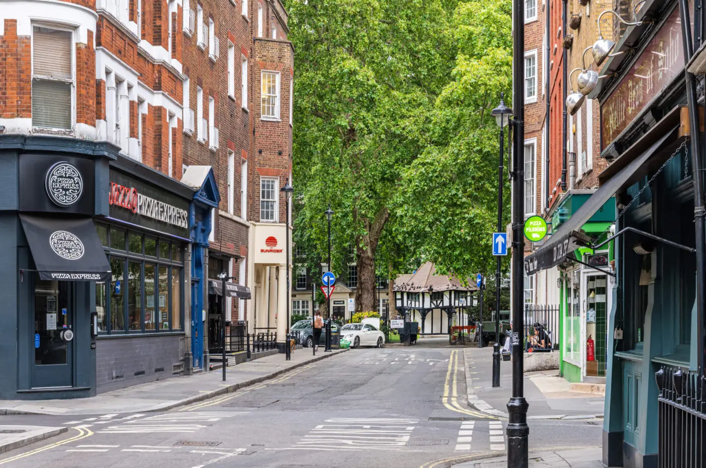 A quiet city street with various shops, including a pizza restaurant and a pub. The street is lined with brick buildings and has a few parked cars. A large green tree, reminiscent of the peaceful ambiance near The Resident Soho, adds a touch of nature to the urban setting.