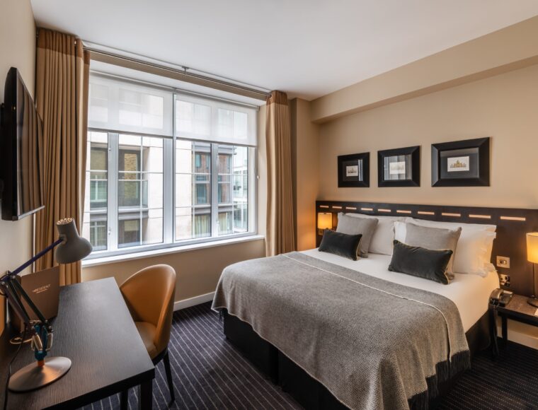 A modern hotel room at The Resident Victoria features a large bed with grey and white bedding, flanked by two nightstands with lamps. A desk with a chair and a wall-mounted TV are positioned on the opposite wall. A large window allows ample natural light to fill the room, which has a warm, neutral colour palette.