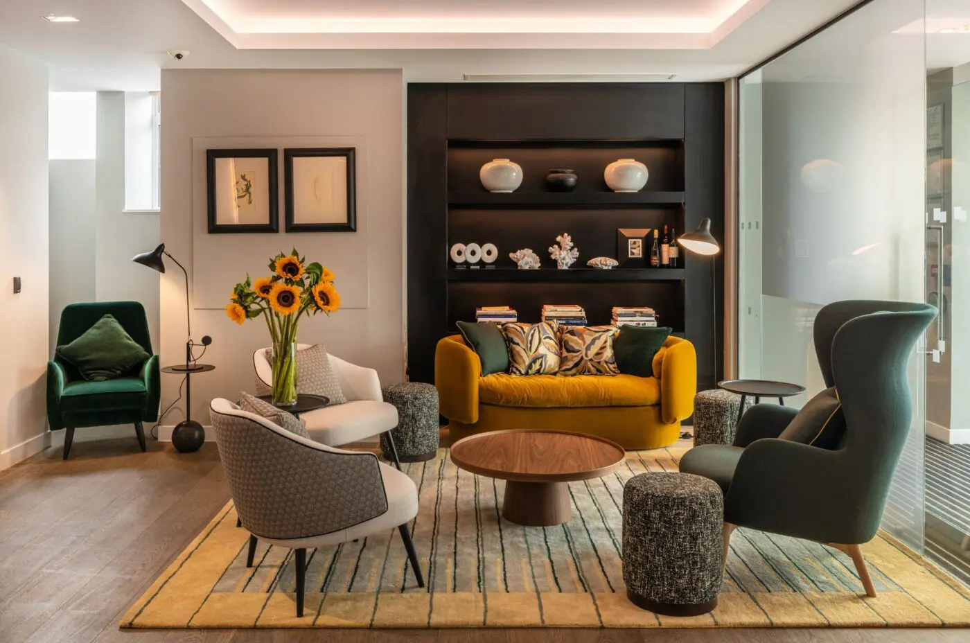 A modern living room with a mustard sofa, two gray chairs, and a green armchair. A wooden coffee table sits on a striped rug. Sunflowers are in a vase, and a shelf with decorative items is in the background. A floor lamp and artwork are on the left wall.