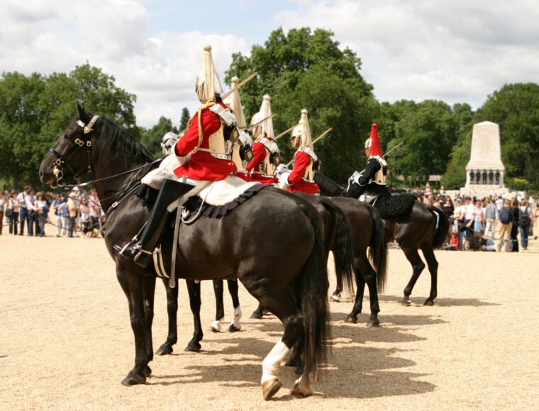 Horse guards in red and white uniforms sit on black horses in a ceremonial formation during the Trooping of the Colour. A crowd observes in the background, with green trees and a stone monument visible under a partly cloudy sky.