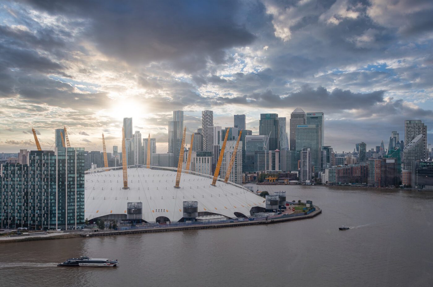The O2 Arena in London sits beside the River Thames, with boats on the water and the modern skyscrapers of Canary Wharf in the background beneath a dramatic sunset sky during the vibrant Greenwich Docklands Festival.