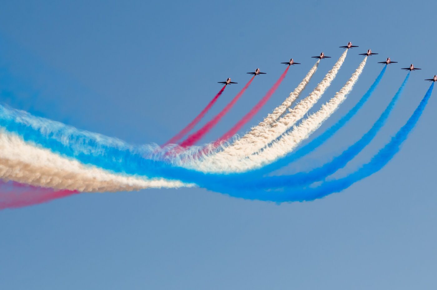 Seven aircraft fly in formation against a clear blue sky at the Southport AirShow, leaving colorful red, white, and blue smoke trails curving gracefully behind them.