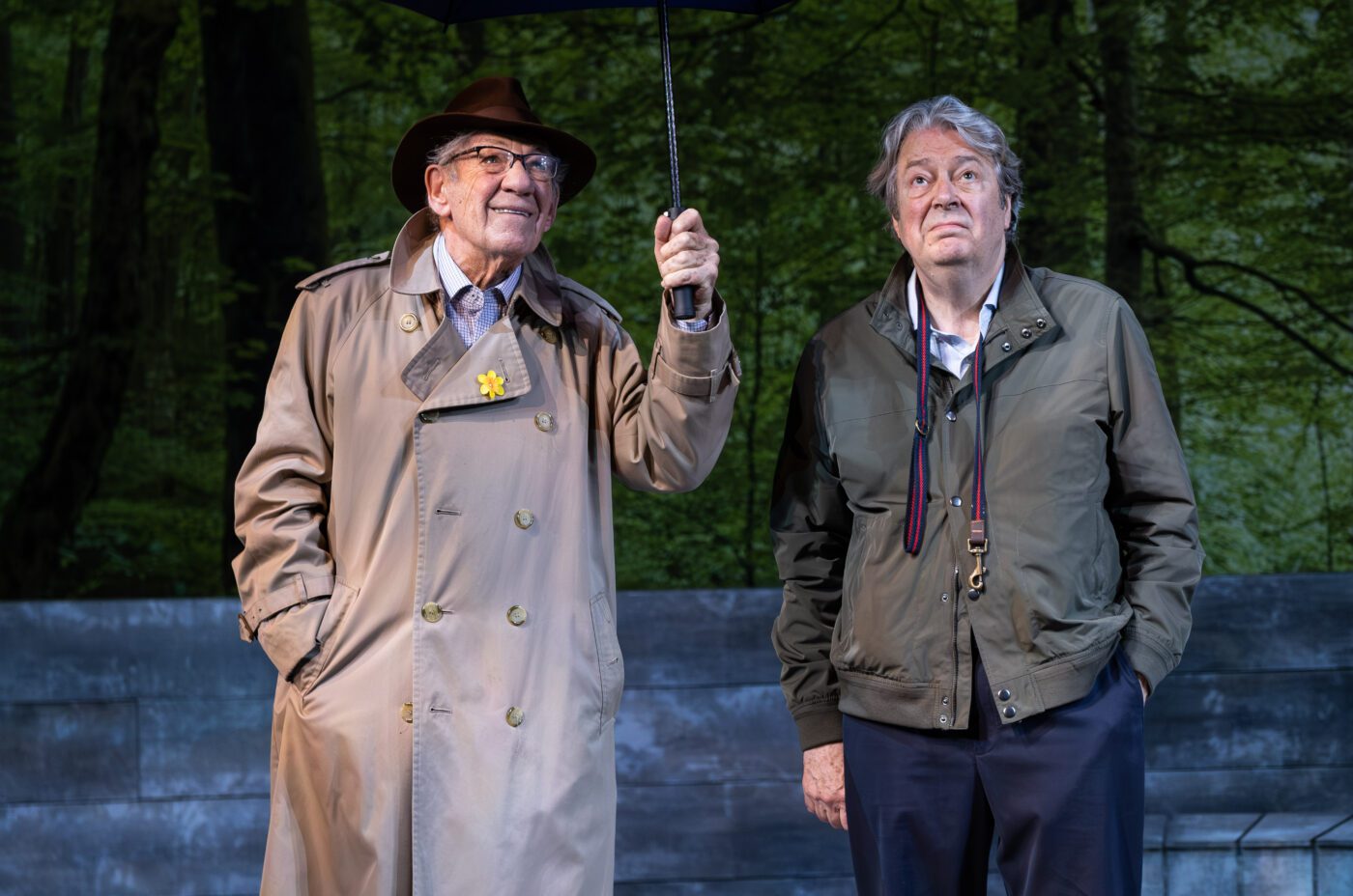 Ian Mckellen and Roger Allam on stage, sheltering from the rain under a shared umbrella.