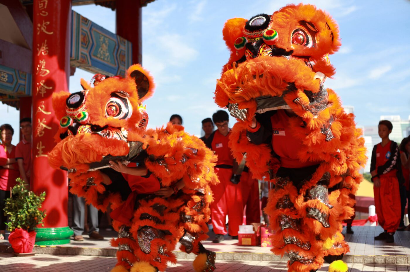 Two vibrant orange lion dance costumes are worn by performers during a Chinese New Year celebration outdoors, with onlookers and decorative red pillars in the background under a sunny sky.