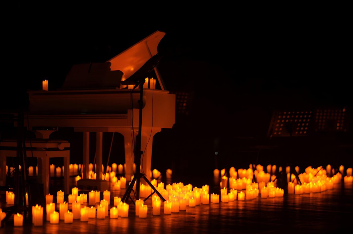 A white grand piano on a dimly lit stage, surrounded by numerous glowing candles, creates a warm and intimate atmosphere—perfect for an International Beatles Week tribute. Music stands are visible in the background.