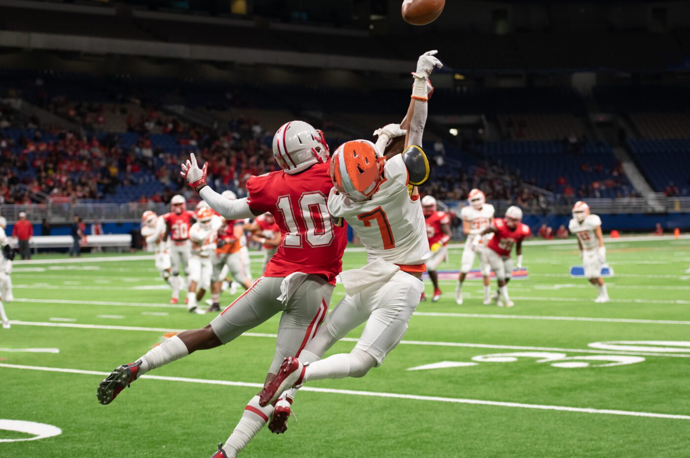 A football player in a white uniform leaps to catch a pass while being closely defended by a player in red on the green Kew Fete field in a stadium, with other players and fans visible in the background.