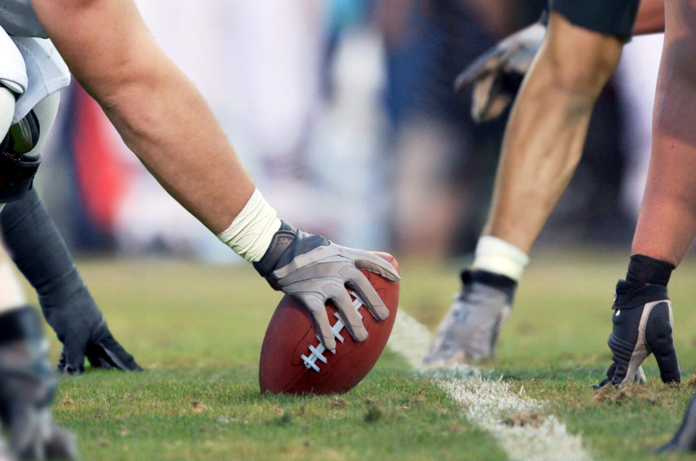 Close-up of American football players at the line of scrimmage, one player's gloved hand gripping the football, ready to snap, evoking the competitive spirit found at events like Kew Fete, with others poised for action on a grassy field.