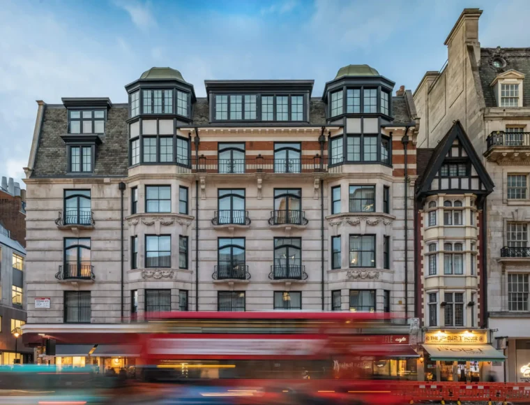A wide shot of an ornate, multi-story building with bay windows and balconies, set on a busy city street. A red double-decker bus passes in front, creating a motion blur effect. Other historic buildings line the street.