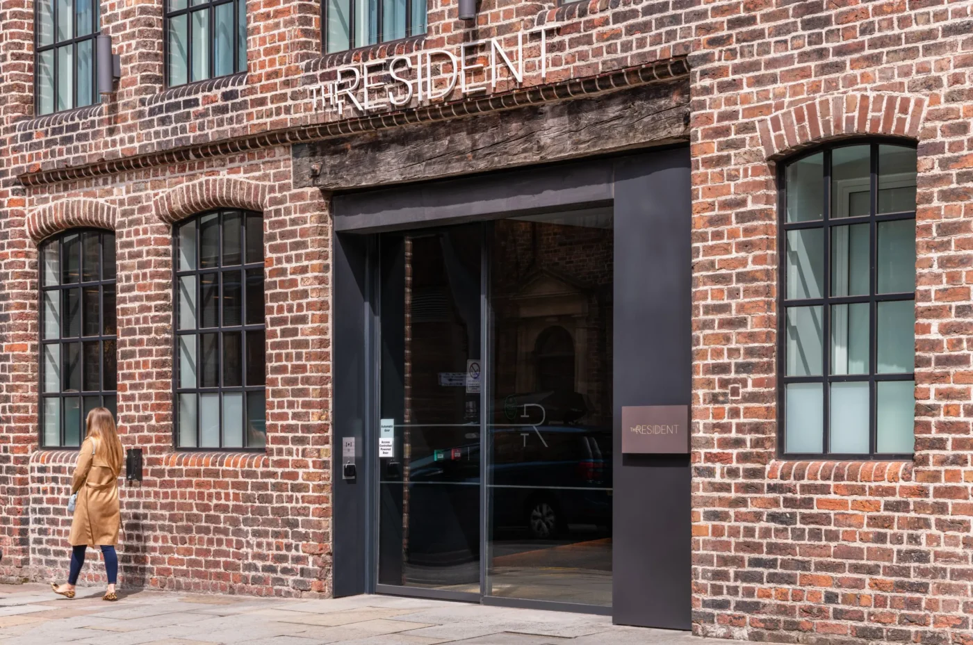 A woman in a tan coat walks past a brick building with black-framed windows and a large glass door. Above the entrance, metal letters spell out 