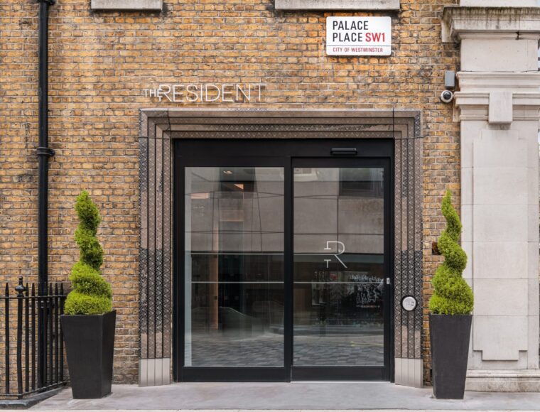 Glass entrance door of The Resident hotel with two spiral-shaped potted topiary plants on either side, set in a brick building. A street sign above reads “Palace Place SW1, City of Westminster.”.