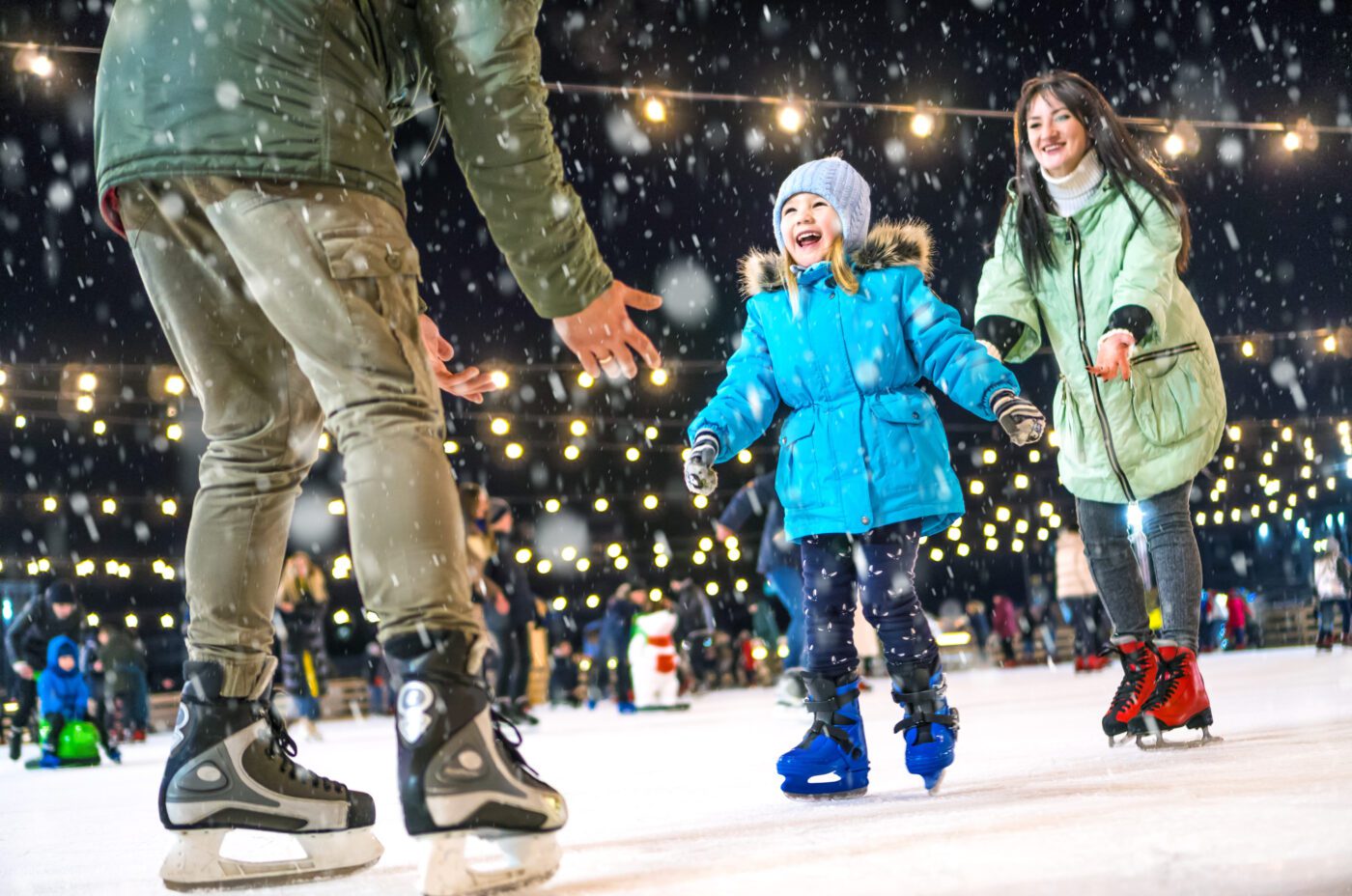A joyful family ice skating outdoors in the evening under string lights at the Christmas Market. A child in a blue coat and hat skates between an adult's outstretched arms and another adult, all smiling amidst gently falling snow.