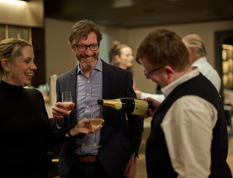 A small group of people is gathered at an indoor event. A man is pouring champagne into glasses held by a woman and another man. They are all smiling and appear to be enjoying a social gathering.