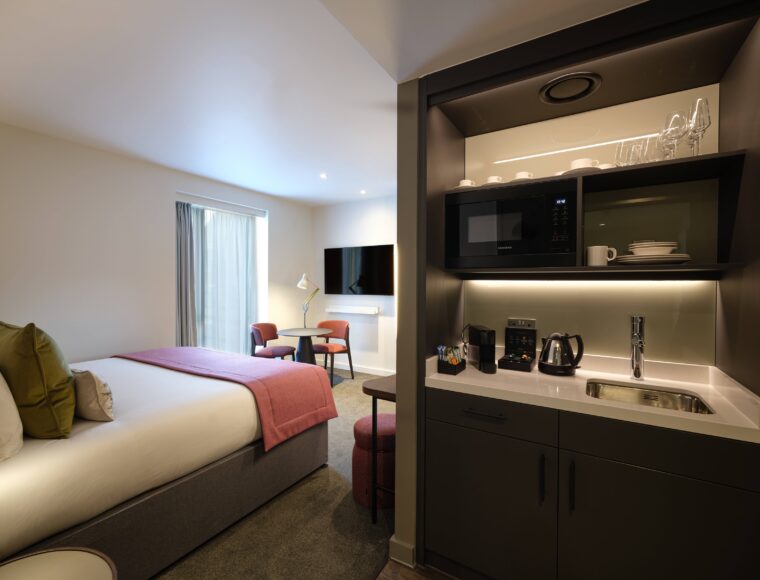 Modern hotel room with a large bed, pink throw blanket, and green pillows. Includes a kitchenette with a microwave, coffee maker, and sink. There's a flat-screen TV on the wall, a small round table, chairs, and a window with curtains.