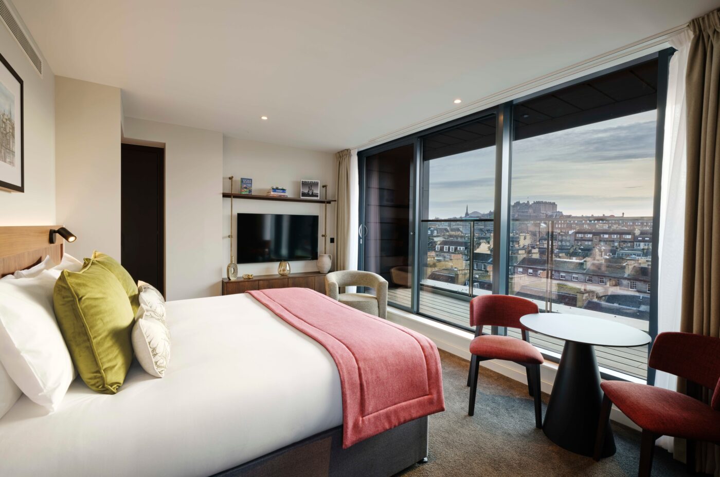 Hotel room with a large bed, red throw blanket, and decorative pillows. There's a TV on the wall, two red chairs, and a table by a large window showcasing a cityscape. Natural light streams in, highlighting the modern, cozy decor.