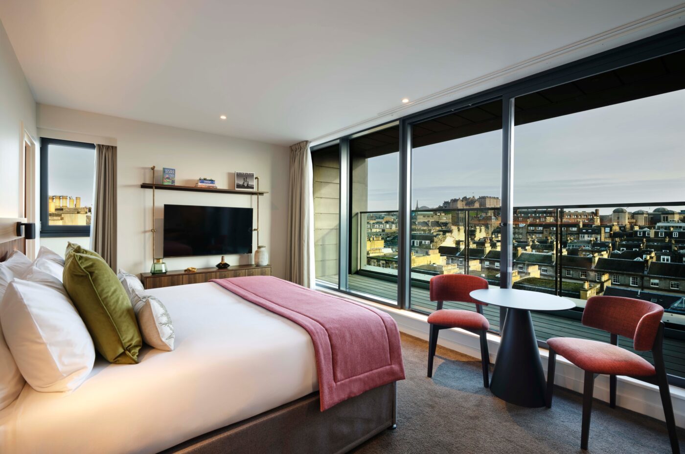 A modern hotel room with a king-sized bed adorned with white sheets and pink accents. A wall-mounted TV faces the bed. Large windows showcase a cityscape view with buildings under a cloudy sky. Two chairs and a small round table sit nearby.