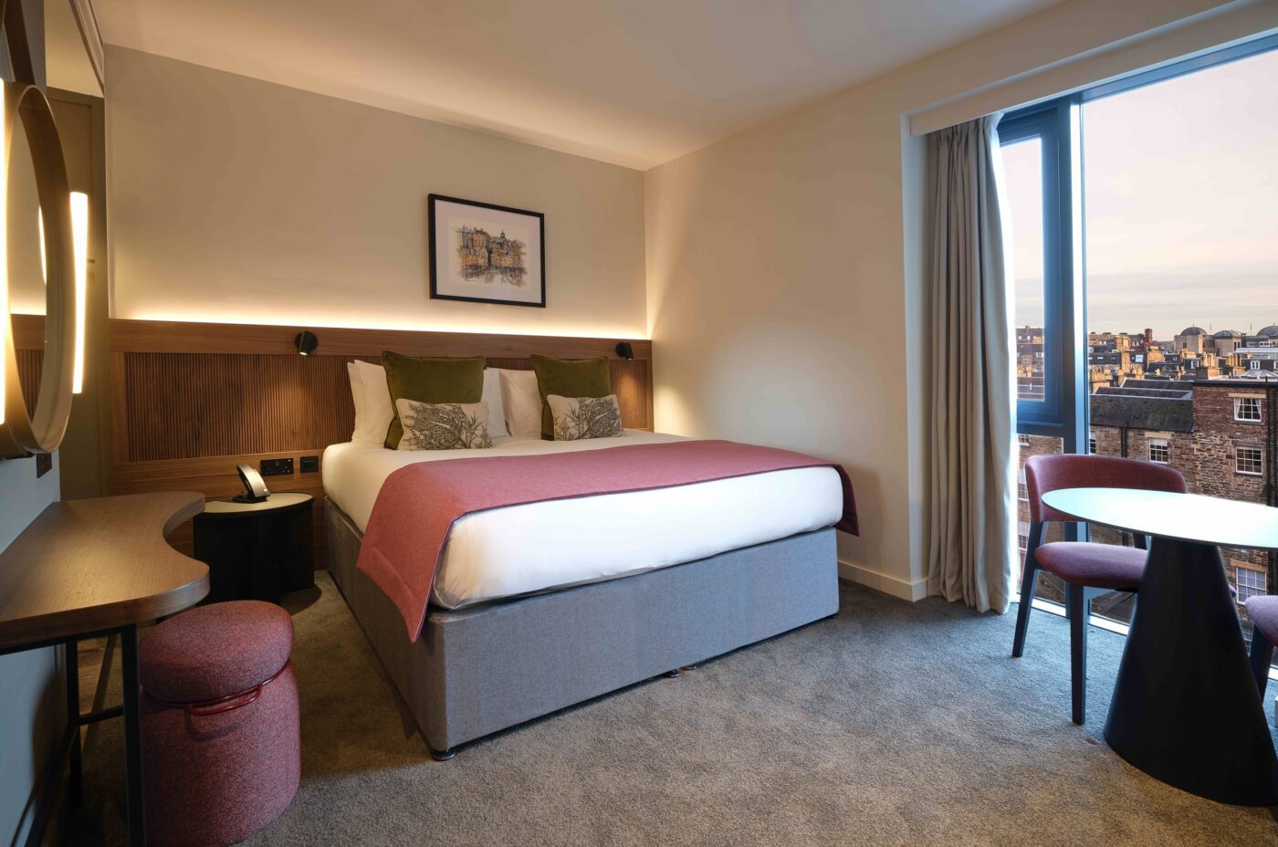 A cozy hotel room with a large bed adorned with white and red bedding. The room features a wooden headboard, a wall lamp, a circular mirror, and a round table with two chairs by a window offering a view of a cityscape.