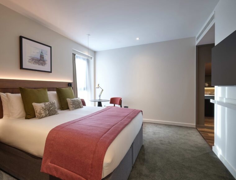A modern hotel room featuring a large bed with white linens, green and white pillows, and a red blanket. A wooden headboard adorns the bed. There's a desk with a lamp, a chair, and a wall-mounted art piece. The room has soft lighting and carpeted flooring.