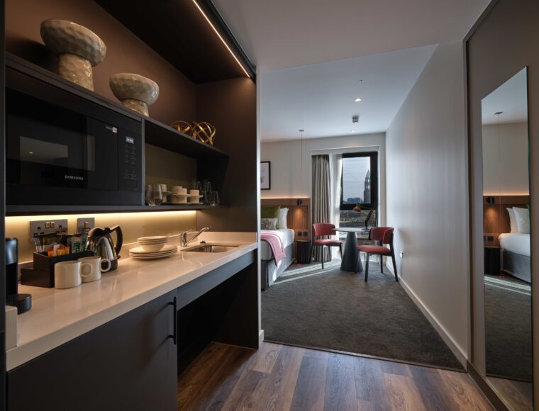 A modern hotel suite with a kitchenette on the left featuring a microwave, sink, and neatly arranged dishes. In the background, a cozy bedroom with a neatly made bed, armchairs, a table, and a large window with a city view.