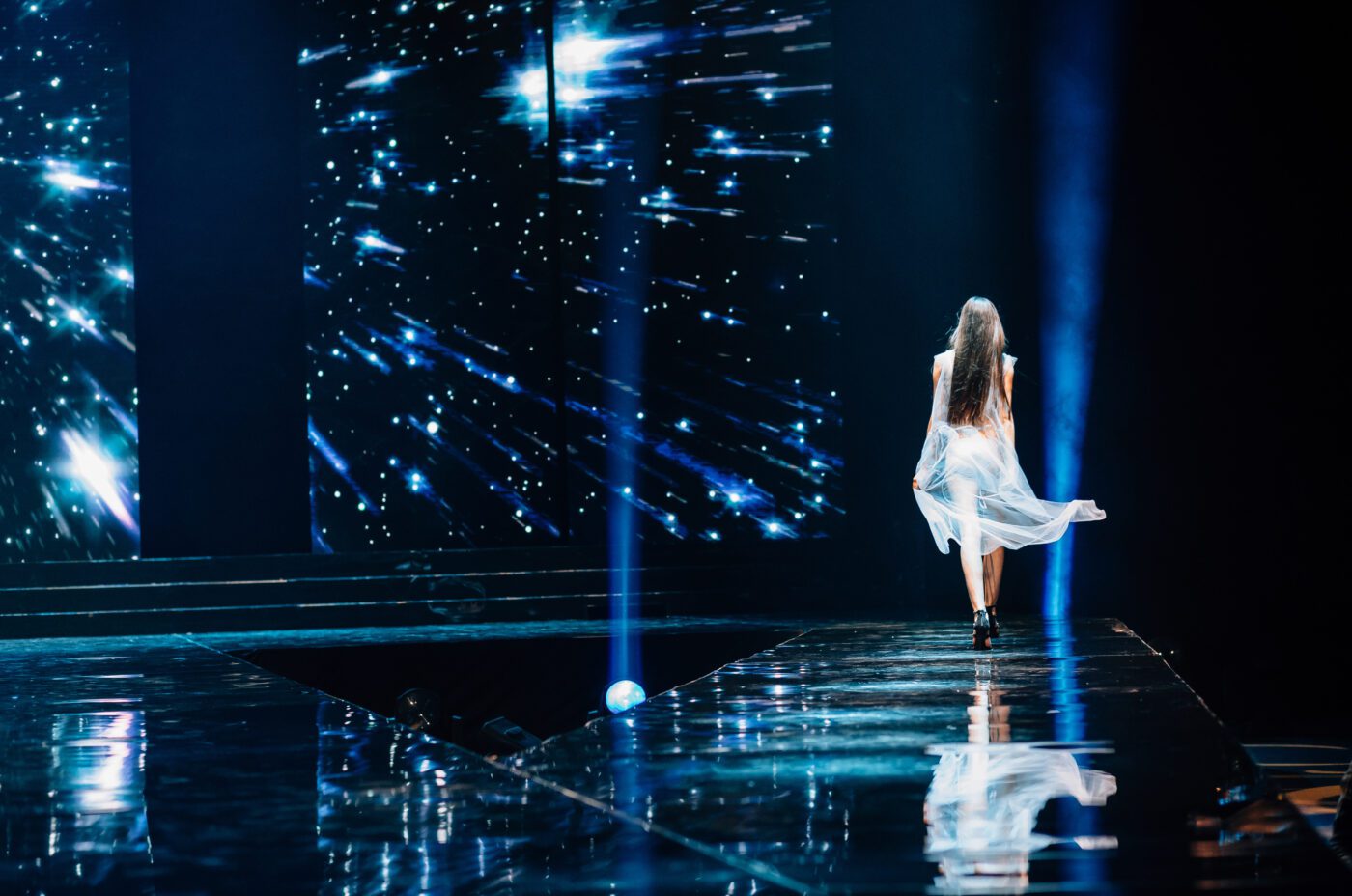 A female model in a flowing white dress walks down a glossy runway. The background displays a starry light effect, creating a cosmic atmosphere. The stage is illuminated by blue spotlights.