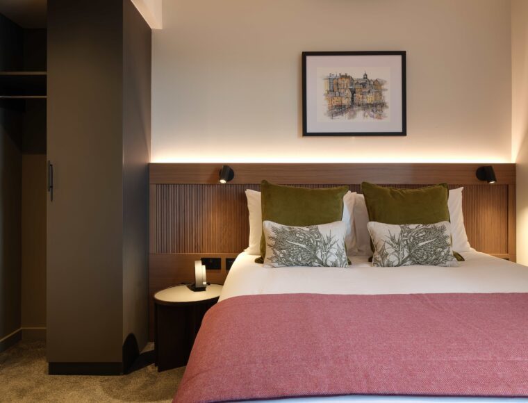 A cozy hotel room with a neatly made double bed covered in white and pink linens, green pillows, and decorative cushions. A framed artwork hangs above the bed. A small round bedside table with a lamp is on the side. The room has warm ambient lighting.