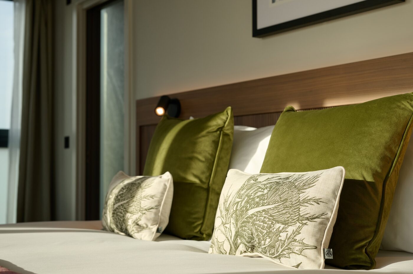 A cozy bedroom scene featuring a bed with two large green velvet pillows and two smaller decorative pillows with leaf patterns. Soft sunlight filters through a window, casting warm light on the bed's wooden headboard and white sheets.