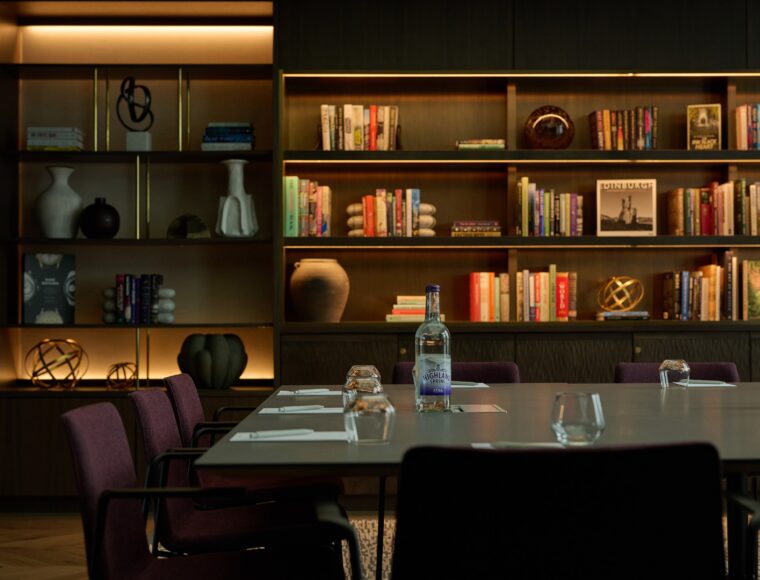 A modern conference room with a long table and chairs. The background features wooden bookshelves filled with books, decorative objects, and subtle warm lighting. A bottle and glasses are set on the table, adding sophistication to the setting.