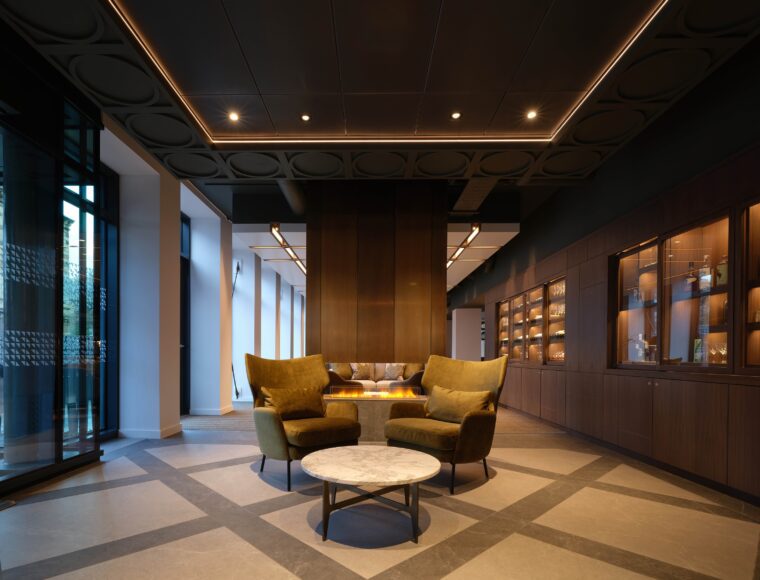 A modern, elegant lounge area with two velvet armchairs and a round marble table in front of a sleek fireplace. The room features large windows, wooden shelving with warm lighting, and a geometric-patterned floor.