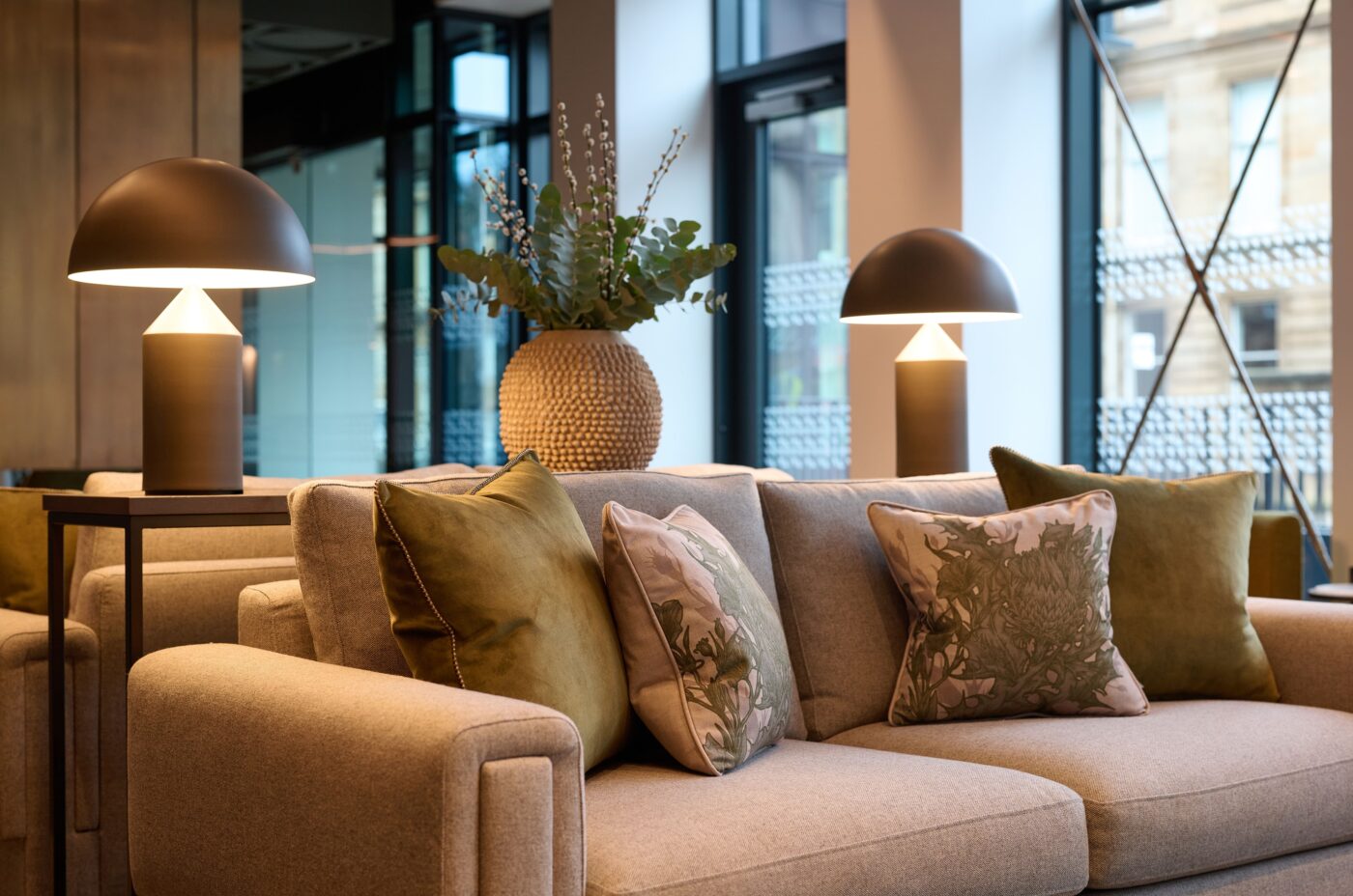 A modern living room with a beige sofa adorned with green and floral cushions. Two matching lamps with dome-shaped shades stand on either side. A large woven vase with leafy plants decorates the background. Large windows allow natural light in.