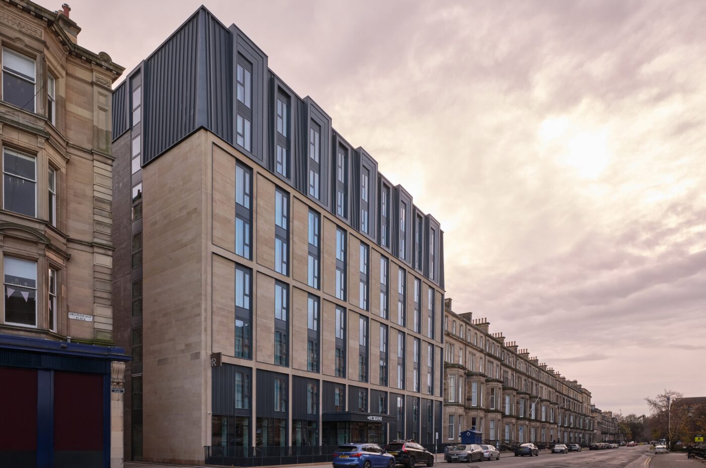 A street view of a modern, multi-story building with large windows, this architectural marvel harmonizes stone and metal cladding. Nestled alongside traditional stone structures under a partly cloudy sky, it stands proudly near the vibrant venues of the Edinburgh International Festival.