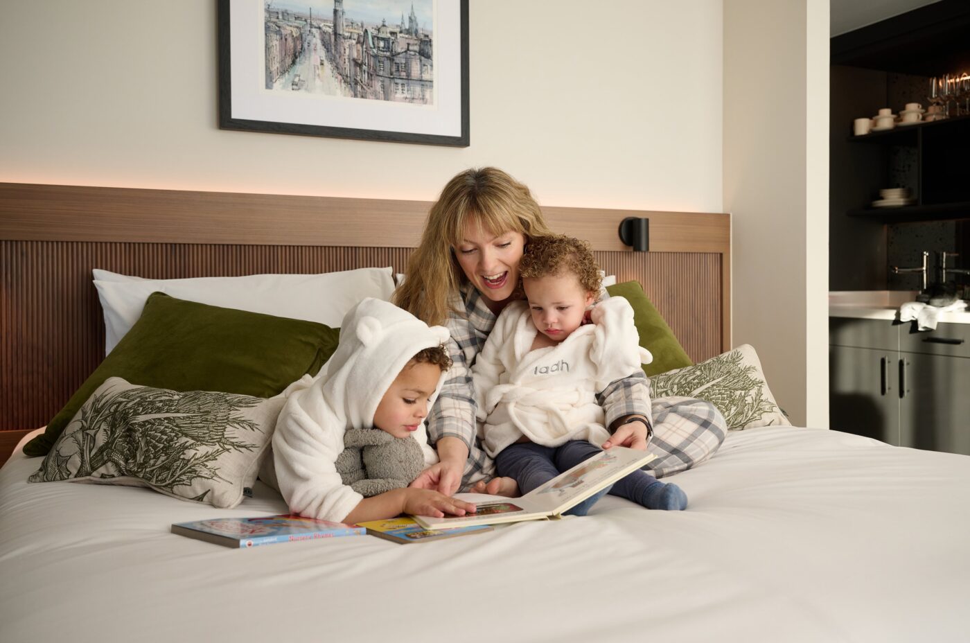 A woman and two young children sit on a bed in a cozy setting. The woman is reading a book to the children, who are wearing hooded robes. There are framed pictures on the wall and plush green pillows on the bed.