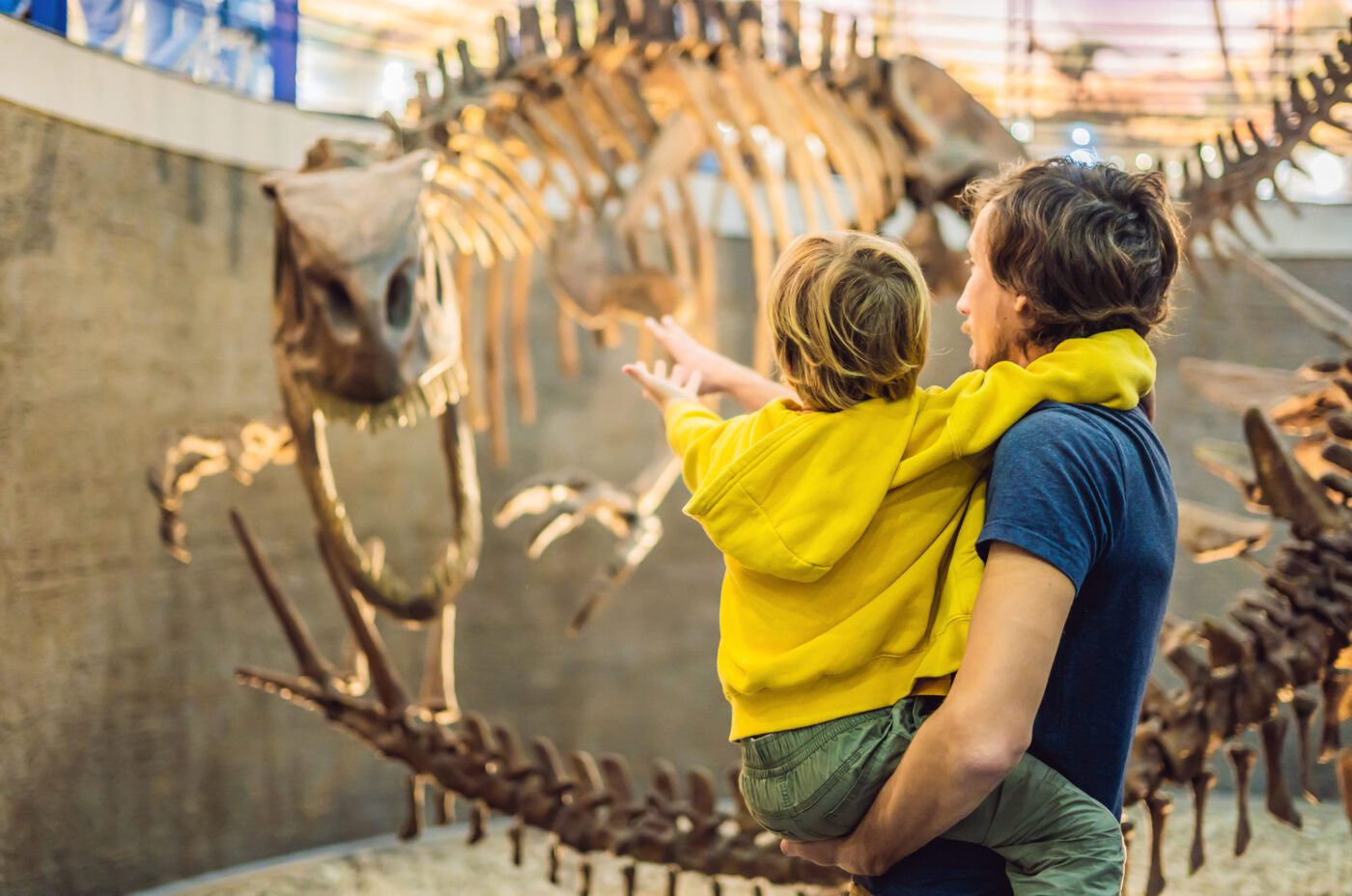 At the Open Doors Festival, a man holds a child in a yellow hoodie as they both marvel and point at a large dinosaur skeleton displayed in the museum. The exhibit area, with its glass ceiling, creates an engaging and educational atmosphere that captivates visitors of all ages.
