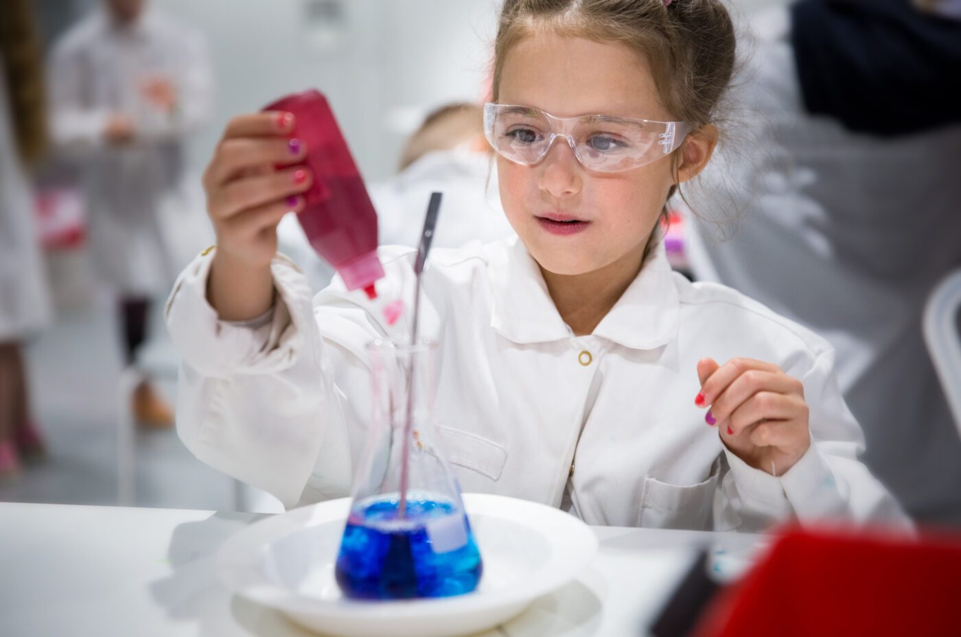 A young girl in safety goggles and a lab coat, participating in the Open Doors Festival, carefully adds red liquid from a dropper to a beaker of blue liquid. Her focus and engagement shine as she demonstrates a science experiment in the lively laboratory setting.