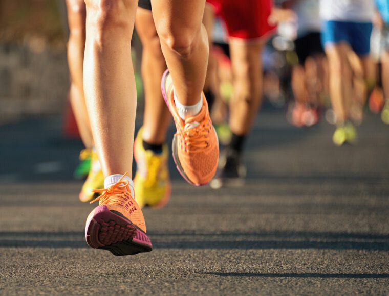 Close-up of runners' legs and colorful sneakers during the London Marathon. Athletes are in motion, their speed captured by a blurred background filled with participants. The focus is on the foreground runner’s bright orange shoes, vibrant against the iconic city's backdrop.