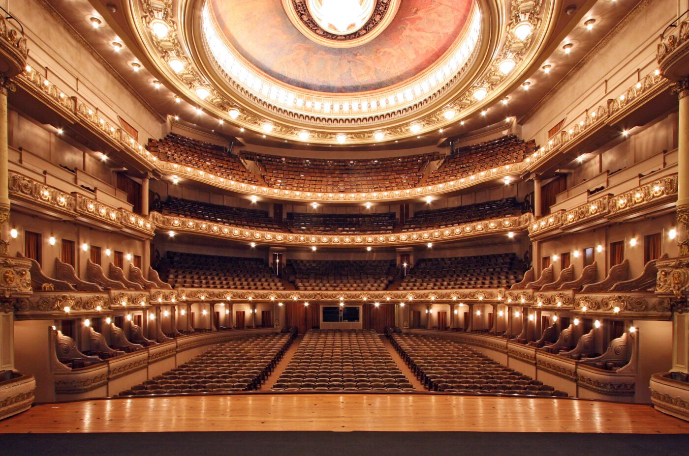 View from the stage of an ornate, empty theater at the V&A Museum, with multiple balconies, intricate gold detailing, and rows of seats facing the stage, illuminated by warm lighting and a large, circular ceiling mural.