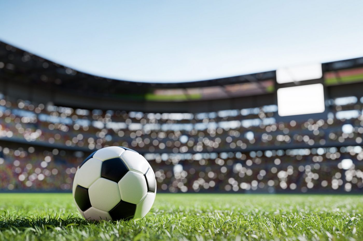 A black and white soccer ball rests on a lush green field inside a large, brightly lit stadium. The stands are blurred in the background, filled with colorful seats and illuminated by sunlight.