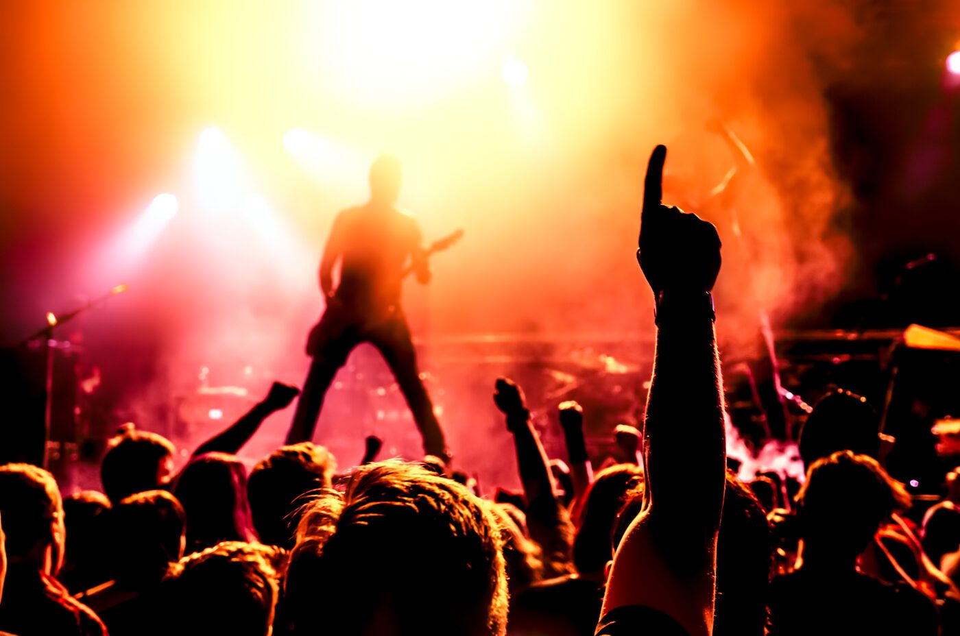 A silhouette of Bruce Springsteen performing amid vibrant lights and smoke during International Beatles Week. In the foreground, a cheering crowd raises their arms, with one hand prominently pointing upward. The scene captures the energetic atmosphere of this lively tribute concert.