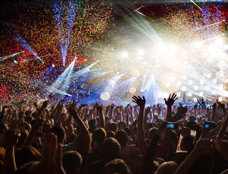 A vibrant concert scene during International Beatles Week features a crowd raising their hands under colorful confetti. Bright stage lights illuminate the area, creating an energetic atmosphere as fans capture the moment with their phones.